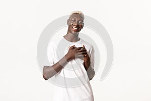 Portrait of happy and touched african american blond man, smiling and holding hands on heart delighted, standing white