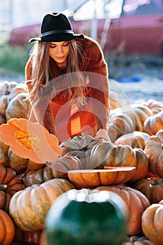 Portrait of happy woman choosing ripe orange pumpkin on farmers market in brown sweater, dress. Cozy autumn vibes