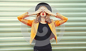 Portrait happy woman covering her eyes with her hands making wish wearing yellow jacket, black hat on metal wall