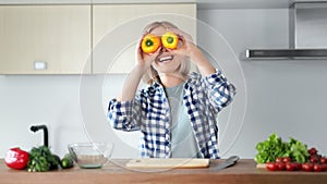 Portrait of happy woman dancing having fun with yellow peppers on eyes at home kitchen