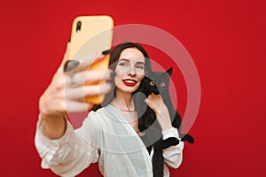 Portrait of happy woman in light clothing taking selfie with black cat on smartphone, hugging pet and looking into smartphone