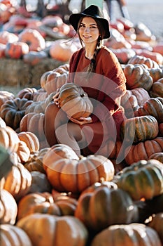 Portrait of happy woman with ripe orange pumpkin in hands on background of farmers market in brown sweater and hat. Cozy