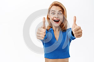Portrait of happy woman showing thumbs up with amazed face, say yes, approve smth awesome, stands over white background