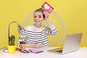 Portrait of happy woman sitting at workplace holding heart icon over head, recommending to follow.