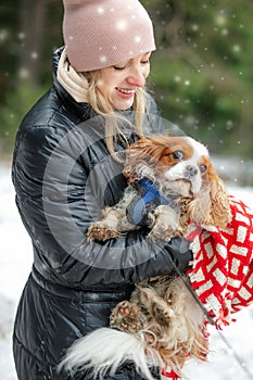 Portrait of happy woman and spaniel dog in winter forest. Woman hold in hands cute spaniel puppy. Walking in snowfall.