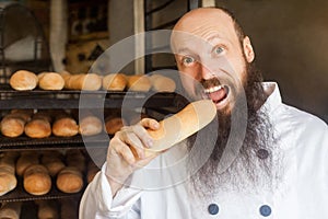 Portrait of happy young adult hungry baker with long beard in white uniform standing in his workplace and tasty fresh baked long