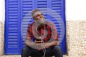 Happy young african man sitting outdoors with mobile phone and looking away