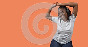 Portrait of happy young African woman dancing and listening to music with headphones, arms up, dressed in white t-shirt and blue