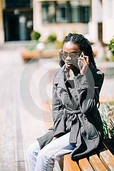 Portrait of happy young african woman sitting outside on bench reading a text message on her mobile phone