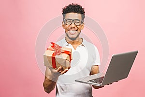 Portrait of happy young afroamerican man using laptop comruter, gift box  and checkin isolated against pink photo