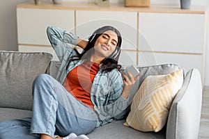 Portrait of happy young Arab woman in wireless headphones relaxing on couch, listening to music on smartphone at home