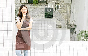 Portrait of a happy young Asian barista in apron looking away and thinking in front of her small coffee shop, waist up, copy space