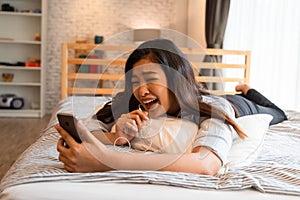 Portrait of happy young Asian girl in casual clothing lying down on bed while making a video call with smartphone in