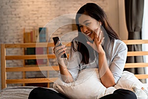 Portrait of happy young Asian girl in casual clothing sitting on bed while making a video call with smartphone in