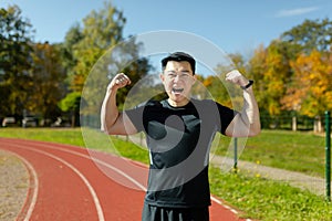 Portrait of a happy young Asian male athlete standing in a stadium, rejoicing at the camera for winning and showing the