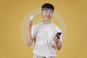 Portrait of happy young asian man holding smartphone and credit card for shopping online isolated on yellow background