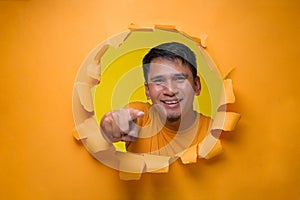 Portrait of Happy young Asian man pointing forward, looking at camera, poses through torn paper hole