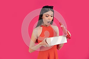 Portrait of a happy young asian woman shocked open gift box isolated over pink background. dressed in orange dress and sunglasses