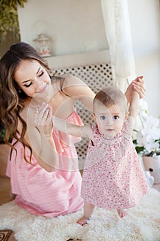 Portrait of happy young attractive mother playing with her baby girl near window in interior at haome. Pink dresses on mother and