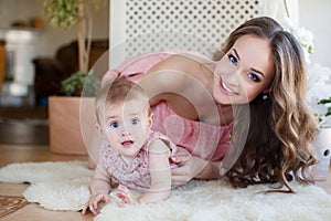 Portrait of happy young attractive mother playing with her baby girl near window in interior at haome. Pink dresses on mother and