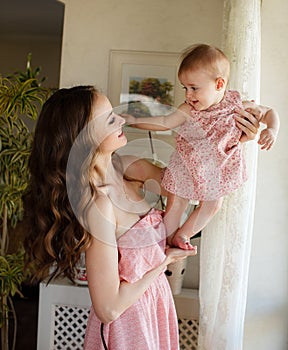 Portrait of happy young attractive mother playing with her baby girl near window in interior at haome. Pink dresses on mother and