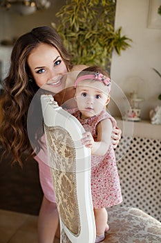 Portrait of happy young attractive mother playing with her baby girl near window in interior at haome. Pink dresses on mother and