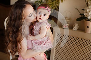 Portrait of happy young attractive mother playing with her baby girl near window in interior at haome. Pink dresses on mother and