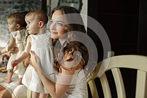 Portrait of happy young attractive mother playing with her baby girl near window in interior at haome. Pink dresses on mother and