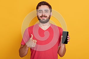 Portrait of happy young bearded hipster man wearing red casual t shirt and showing ok gesture, holding phone with blank screen,