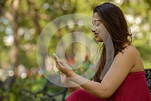 Portrait of happy young beautiful Indian woman using phone at the park