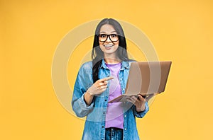 Portrait of happy young beautiful surprised woman with glasses standing with laptop isolated on yellow background.