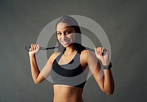 Portrait of happy young biracial woman in sports bra holding jump rope behind neck in studio