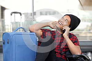 Happy young black man sitting on floor at station with travel bags and talking with mobile phone