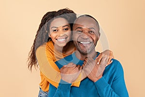 Portrait of happy young black woman hugging her middle aged husband from behind over peach studio background