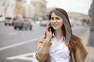 Portrait of happy young brunette woman in beige coat talking on