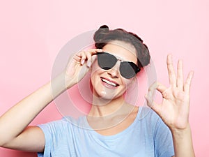 Portrait of a happy young brunette woman wearing sunglasses showing ok gesture over pink background