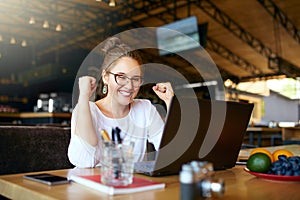 Portrait of happy young business woman celebrating success with arms up in front of laptop. Mixed race female won a lot