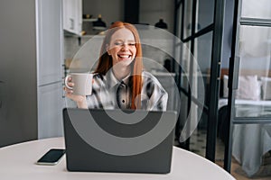 Portrait of happy young business woman remote working or studying on laptop computer sitting at table, holding in hand
