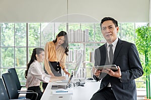 Portrait of happy young businessman holding tablet and looking at camera while his colleagues working in office background