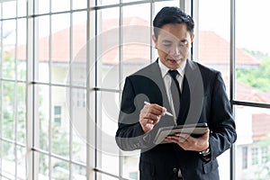 Portrait of happy young businessman looking at business document in tablet, standing near the window in office background