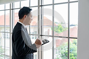 Portrait of happy young businessman looking at business document in tablet, standing near the window in office background