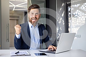 Portrait of a happy young businessman showing a hand gesture of success and victory to the camera. He sits in the office