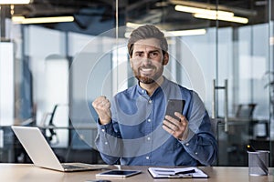 Portrait of a happy young businessman sitting at a desk in the office and looking at the camera, holding a mobile phone