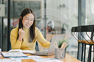 Portrait of a happy young businesswoman celebrating success with arms raised in front of a laptop, fists clenched. The