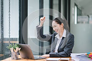 Portrait of a happy young businesswoman celebrating success with arms raised in front of a laptop, fists clenched. The