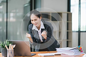 Portrait of a happy young businesswoman celebrating success with arms raised in front of a laptop, fists clenched. The