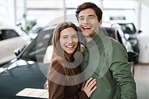 Portrait of happy young Caucasian couple posing and smiling at camera near new auto at dealership