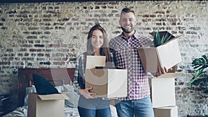 Portrait of happy young couple standing in new house, holding carton boxes, smiling and looking at camera. Family