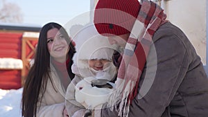 Portrait of happy young family, father, mother and adorable son in warm clothes hugging white rabbit standing in yard on