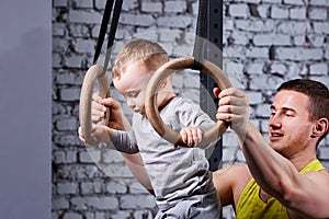 Portrait of the happy young father trains the little son with gimnastic rings against brick wall in the gym.